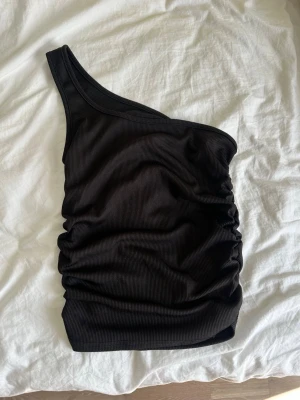 Svart oneshoulder ribbad topp - Snygg svart ribbad topp med oneshoulder-design och rynkade sidor. Perfekt för dig som vill ha en trendig och stilren look. Toppens material är stretchigt och sitter tight mot kroppen.
