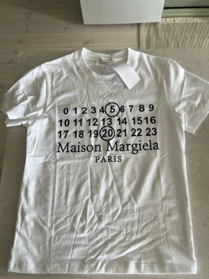 Maison Margiela vit t-shirt med tryck - Maison Margiela t-shirt i vitt med ikoniskt svart tryck framtill med siffror och logga. Klassisk rund hals och korta ärmar. Tillverkad i mjuk bomull för en skön och avslappnad look. Perfekt för dig som gillar minimalistisk och trendig design.