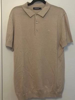 Beige pikétröja från J.Lindeberg - Klassisk beige pikétröja från J.Lindeberg med korta ärmar och krage. Tröjan har två knappar framtill och diskret logga på bröstet. Tillverkad i mjuk bomull som känns skön mot huden. Perfekt till både jeans och shorts.