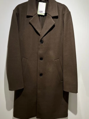 Brun ullkappa med knappar - HM - Stilren brun kappa i ull med klassisk krage och tre svarta knappar framtill. Kappan har två snedställda fickor och en rak passform som ger en tidlös look. Perfekt för kyligare dagar och enkel att matcha med olika outfits.