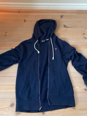 Marinblå hoodie från Levi's med dragkedja - Snygg marinblå hoodie från Levi's i standard fit med dragkedja framtill, två fickor och vit dragsko i huvan. Klassisk liten Levi's-logga på bröstet. Perfekt för en avslappnad och cool stil.