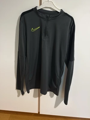 Mörkgrå Nike träningströja - Mörkgrå långärmad träningströja från Nike med halv dragkedja framtill och gulgrönt Nike-märke på bröstet. Tröjan har svarta axelpartier och är tillverkad i ett stretchigt funktionsmaterial som passar perfekt för sport och träning.