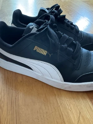 Svarta Puma sneakers med vit sula - Snygga svarta sneakers från Puma med klassisk vit sula och vit sidostripes. Skorna har svarta skosnören, perforerad tå och guldfärgad Puma-logga på sidan. Tillverkade i syntetmaterial och har en sportig, stilren look som passar till det mesta. I väldigt bra skick och rengjorda!