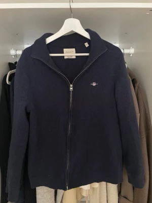 Mörkblå ziptröja från Gant (lite krympt) - Mörkblå ziptröja från Gant med liten broderad logga på bröstet. Tröjan har hög krage, hel dragkedja framtill och är tillverkad i mjukt material. Perfekt för lager på lager och enkel att matcha med olika outfits. Lite krympt.