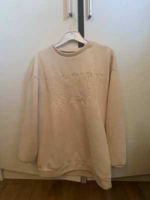 Beige sweatshirt med MALIBU-tryck - Oversized beige sweatshirt med broderat MALIBU-tryck framtill. Tröjan har rund halsringning, långa ärmar och ribbade muddar vid ärmslut och nederkant. Perfekt för en avslappnad och trendig look.