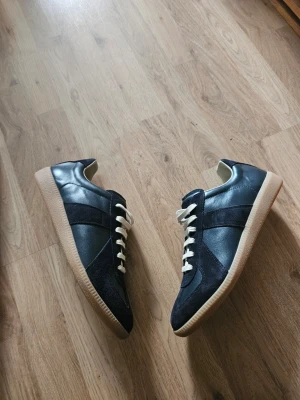Maison Margiela Navy Gats - Snygga mörkblå Maison Margiela Gat sneakers med både mocka och läderpaneler, vita snören och klassisk gummisula. Skorna har en stilren och tidlös design som passar perfekt till jeans eller chinos. Perfekta för dig som gillar en clean och sportig look.