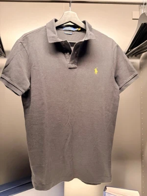 Ralph Lauren - Klassisk svart pikétröja från Ralph Lauren med gul broderad logga på bröstet. Modellen är custom slim fit och har korta ärmar samt krage med knappar. Tillverkad i mjuk bomull som känns skön mot huden.