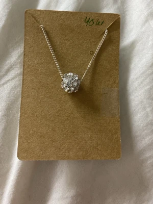 Silverhalsband med glittrande pärlor - Snyggt halsband i silverfärg med en rund pärlformad hängberlock täckt av små gnistrande stenar. Kedjan är tunn och elegant, perfekt för att ge en extra touch till din outfit. Passar dig som gillar stilrena och glittriga accessoarer.