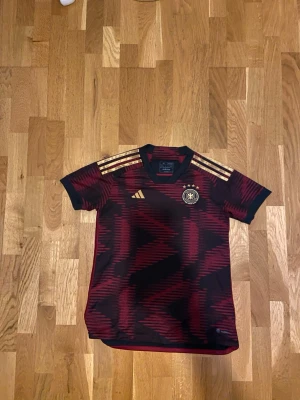 Tysklands matchtröja Adidas röd/svart - Säljer en officiell Tyskland matchtröja från Adidas i rött och svart med guldiga detaljer och tre ränder på axlarna. Tröjan har korta ärmar, V-ringning och Tysklands landslagsemblem på bröstet. Perfekt för fotbollsfans! Den är äkta direkt från Tyskland