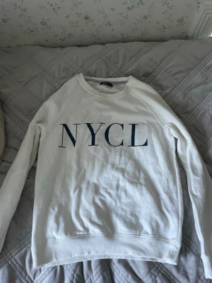Vit sweatshirt från Gina Tricot - Säljer en vit sweatshirt från Gina Tricot med trycket 'NYCL' i mörkblå bokstäver på bröstet. Tröjan har rund halsringning, långa ärmar och mjukt material på insidan. Perfekt för en avslappnad och trendig look. Har inte använt den så många gånger så den är fortfarande ganska mjuk på insidan.
