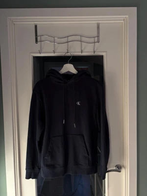 Mörkblå hoodie från Calvin Klein - Mörkblå hoodie från Calvin Klein med liten CK-logga på bröstet. Klassisk modell med huva och dragsnören samt stor magficka framtill. Mjuk och skön bomullsblandning, perfekt för en avslappnad stil.