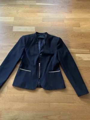 Svart kavaj med dragkedjedetaljer Zara - Stilren svart kavaj från Zara Basic med markerad midja och coola dragkedjor vid fickorna. Kavajen har lång ärm, rak passform och är tillverkad i ett slätt tyg med lätt glans. Perfekt för en trendig och modern look.