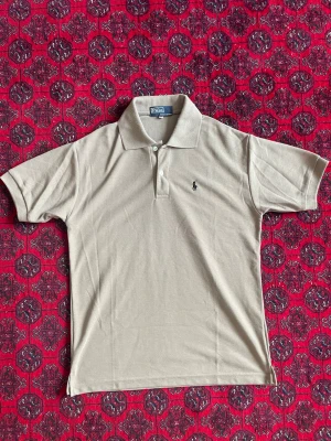 Polo Ralph Lauren Pike - Klassisk beige pikétröja från Polo Ralph Lauren med broderad logga på bröstet. Tröjan har krage, korta ärmar och knäppning framtill. Perfekt för en stilren och avslappnad look.
