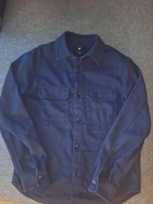 Mörkblå overshirt från H&M - Stilren mörkblå overshirt från H&M med klassisk krage och två stora bröstfickor med lock. Skjortan har knäppning framtill och vid ärmsluten. Tillverkad i ett mjukt, tyg som ger en avslappnad look och bra till alla väder.skriv om du har frågor eller vill se mera bilder ✅