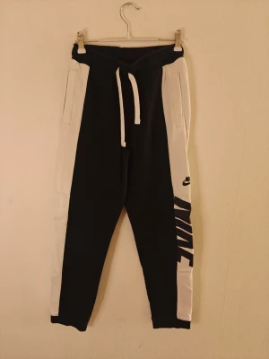 Nike mjukisbyxor  - Säljer ett par snygga mjukisbyxor från Nike i svart och vitt med breda kontrastpaneler och stort Nike-tryck på benet. Byxorna har dragsko i midjan och är gjorda i mjuk bomullsblandning med fickor på sidorna. Perfekta för chill eller träning. Storlek M för barn.