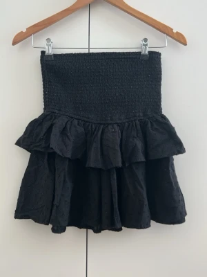 Svart volangkjol med smock - Supersöt svart kortkjol med bred smock i midjan och dubbla volanger. Kjolen har en luftig känsla och är perfekt för dig som gillar en feminin och trendig stil. Materialet känns mjukt och lätt, och volangerna ger extra rörelse till outfiten.