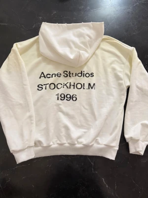 Vit hoodie från Acne Studios - Snygg vit hoodie från Acne Studios med tryck på ryggen där det står 'Acne Studios STOCKHOLM 1996'. Klassisk passform med huva och ribbade muddar. Perfekt för dig som gillar stilrena plagg med en touch av streetwear.