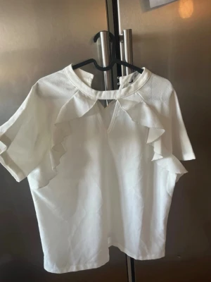 Vit blus med volangdetaljer - Snygg vit blus med korta ärmar och volangdetaljer längs axlarna. Blusen har en liten v-ringning framtill och är gjord i ett mjukt tyg som känns skönt mot huden. Perfekt för dig som gillar stilrena plagg med en twist.