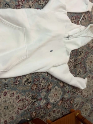 Vit hoodie från Polo Ralph Lauren - Snygg vit hoodie från Polo Ralph Lauren med klassisk huva och dragsko. Har en liten broderad logga på bröstet och stor magficka framtill. Tillverkad i mjukt material som känns riktigt skönt mot huden. Perfekt för dig som gillar stilrena och tidlösa plagg.