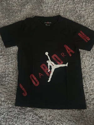 Svart Jordan t-shirt med logga - Säljer en svart t-shirt från Jordan med det klassiska Jumpman-trycket i vitt och texten JORDAN i rött diagonalt över framsidan. T-shirten har rund hals och korta ärmar, perfekt för en sportig och avslappnad stil.