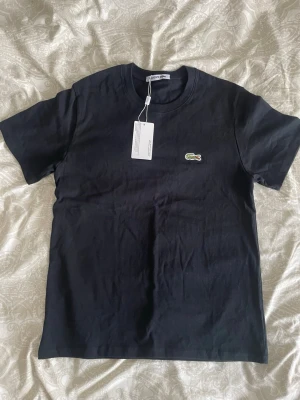 Svart t-shirt från Lacoste - Klassisk svart t-shirt från Lacoste med den ikoniska krokodilloggan broderad på bröstet. T-shirten har rund halsringning och korta ärmar. Tillverkad i mjuk bomull för en skön känsla och enkel stil.