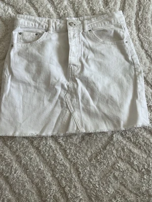 Vit jeanskjol med rå kant från Gina - Snygg vit jeanskjol med klassiska fem fickor och rå kant nertill. Kjolen har knapp och dragkedja framtill samt bälteshällor i midjan. Perfekt till sommaren för en fräsch och enkel look.