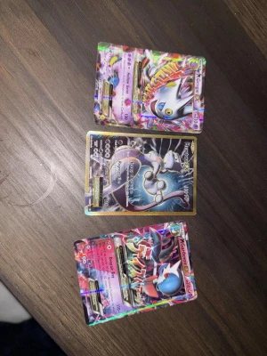 Pokémonkort Mega Mewtwo EX - Tre Pokémonkort med Mega Mewtwo EX-motiv. Två av korten har färgstarka, holografiska illustrationer i rosa, lila och blått, medan ett kort har en svart och guld design. Alla korten har detaljerade illustrationer och är samlarkort från Pokémon Trading Card Game.