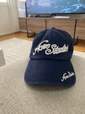 Mörkblå keps från Acne Studios - Snygg mörkblå keps från Acne Studios med vit broderad logga framtill och på skärmen. Klassisk böjd skärm och justerbart spänne bak. Tillverkad i bomullstyg, perfekt för en trendig och avslappnad stil.