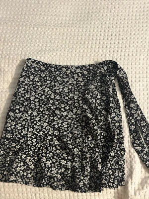 Svart blommig omlottkjol FB Sister - Snygg svart kort omlottkjol från FB Sister med vitt blommönster. Kjolen har volangdetalj nertill och knyts i sidan för en trendig look. Perfekt för dig som gillar en feminin och stilren stil.