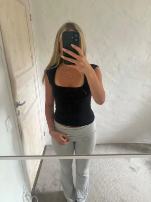 Svart croppad topp med bred urringning - Stilren svart croppad topp med bred urringning och breda axelband. Toppen har en tight passform och är ärmlös, vilket ger en modern och clean look. Perfekt att matcha med höga byxor eller kjol.