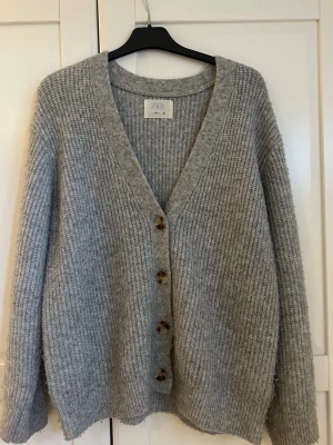 Grå stickad kofta från Zara - Mysig grå kofta från Zara med ribbstickning och djup V-ringning. Koftan har fyra stora knappar framtill och långa ärmar. Är i storlek 164 men skulle säga att den även passar M!! Lite lätt nopprig!❣️❣️