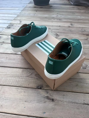 Gröna sneakers, Hammarby  - Stilrena gröna sneakers från Hammarby, Intersport, helt nya. med vit sula och diskreta vita ränder på hälen. Skorna har en låg profil, rund tå och är tillverkade i slätt läder med brunt innerfoder. Perfekta för dig som gillar minimalistisk design och vill sticka ut med färg.
