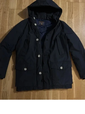 Mörkblå dunjacka från Woolrich - Snygg mörkblå dunjacka från Woolrich med huva och stora fickor framtill. Jackan har silverfärgade knappar och dragkedja, samt ett quiltat innerfoder för extra värme. Perfekt för kalla vinterdagar och har en klassisk, stilren look.