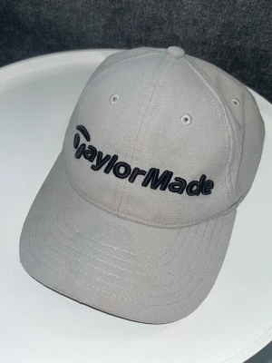 Grå TaylorMade keps - Snygg grå keps från TaylorMade med svart broderad logga framtill. Klassisk böjd skärm och justerbar passform. Tillverkad i slitstarkt tyg, perfekt för golf eller streetwear. One size fits all.