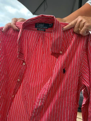 Röd- och vitrandig skjorta Polo Ralph Lauren - Snygg röd skjorta med vita ränder från Polo Ralph Lauren. Klassisk krage, knäppning framtill och broderad logga på bröstet. Skjortan är i bomull och har lång ärm. Perfekt för en stilren look.