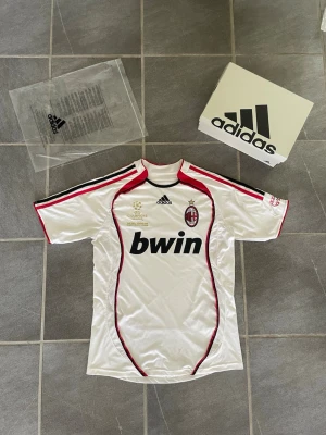 AC Milan fotbollströja Adidas - Säljer en vit AC Milan fotbollströja från Adidas med röda och svarta detaljer. Tröjan har klubbmärke, Champions League-märke och Bwin-tryck på bröstet. Tillverkad i lätt och ventilerande material, perfekt för fotboll eller streetwear.