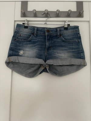 Blå jeansshorts med upprullad kant - Snygga blå jeansshorts med upprullad kant och slitning på ena benet. Klassisk femficksmodell med knapp och dragkedja framtill. Perfekta för varma dagar och ger en avslappnad look.