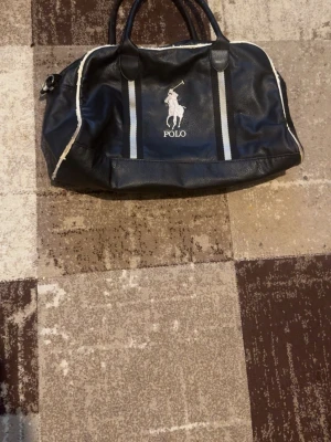 Svart weekendbag från Polo - Snygg svart weekendbag från Polo med vita detaljer och två handtag. Väskan har ett stort huvudfack, vita ränder framtill och en broderad logga med häst. På baksidan finns en ficka med dragkedja och en stor vit siffra 3 på sidan. Materialet är konstläder.