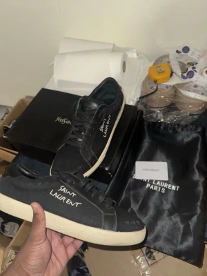 Svarta Saint Laurent sneakers - Svarta sneakers från Saint Laurent med vit sula och handskriven logga på sidan. Klassisk låg modell med svarta skosnören och ovandel i mocka. Perfekta för dig som gillar stilrena och exklusiva skor med en edgy touch.