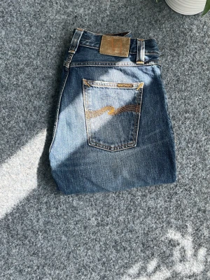 Blå jeans från Nudie Jeans - Säljer ett par klassiska blå jeans från Nudie Jeans med snygg slitning och råa kanter vid bensluten. Jeansen har en rak passform, fem fickor och den ikoniska Nudie-sömmen på bakfickan. Tillverkade i slitstark denim med cool vintagekänsla.