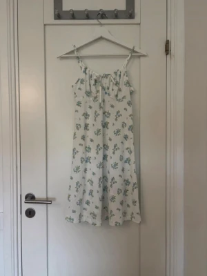 Vit blommig sommarklänning - Söt vit klänning med blå och gröna blommor, smala axelband och knytning fram. Lätt och luftig modell som passar perfekt till varma dagar. Klänningen har en avslappnad passform och slutar strax ovanför knäna.