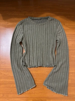 Olivgrön ribbad stickad tröja - Trendig olivgrön stickad tröja med breda ribbor och utsvängda ärmar. Tröjan har en rund halsringning och vågig kant nedtill för en cool look. Perfekt för dig som gillar en avslappnad men ändå stilren vibe.