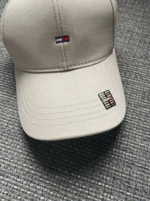 Beige keps från Tommy Hilfiger - Snygg beige keps från Tommy Hilfiger med det klassiska lilla broderade flaggloggan framtill. Kepsen har böjd skärm och justerbart band bak. Tillverkad i bomullstyg för en clean och stilren look.