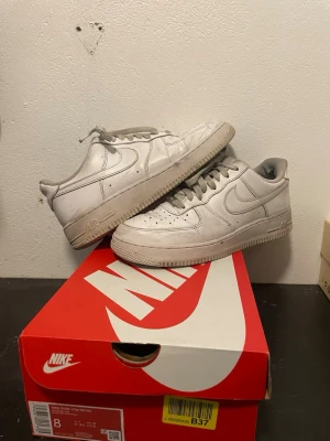 Nike Air Force 1 vita sneakers - Klassiska vita Nike Air Force 1 sneakers i läder med ikonisk swoosh på sidorna och perforerad tå för ventilation. Skorna har en chunky sula och lågt skaft, perfekt för en clean och tidlös streetstil.