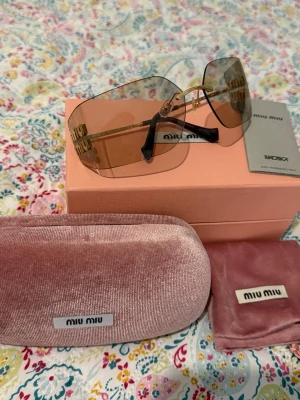 Miu Miu solglasögon med guldton - Säljer ett par trendiga solglasögon från Miu Miu med stora, fyrkantiga linser i ljusbrun ton och guldfärgade detaljer. Glasögonen har en elegant, minimalistisk design och kommer med original rosa sammethölje och putsduk.