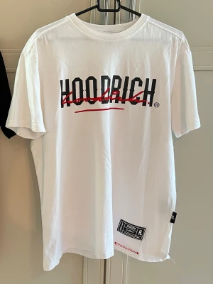 Vit HOODRICH t-shirt med tryck - Säljer en vit t-shirt från HOODRICH med svart och rött tryck både fram och bak. T-shirten har korta ärmar, rund halsringning och detaljer som etikett och röd söm nertill. Perfekt för dig som gillar streetwear och vill sticka ut.