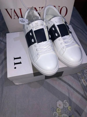 Valentino vita sneakers med blå detalj - Snygga vita sneakers från Valentino med bred mörkblå rem över mitten och vita nitar på sidorna. Skorna har klassisk låg profil, vita skosnören och är tillverkade i läder. Perfekta för dig som gillar stilrena och exklusiva sneakers.