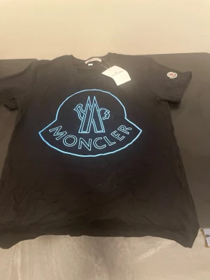 Svart Moncler t-shirt med blå logga - Svart t-shirt från Moncler med stor blå logga framtill och klassisk rund hals. T-shirten har korta ärmar och en Moncler-patch på ena ärmen. Tillverkad i mjuk bomull för en skön känsla och stilren look.