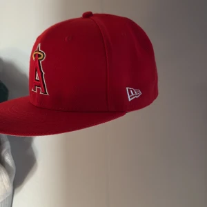 Röd LA Angels keps från New Era - Snygg röd 59FIFTY keps från New Era med LA Angels logga framtill och broderad patch på sidan för 50-årsjubileum. MLB-logga baktill och platt skärm. Tillverkad i 100% polyester, perfekt för dig som gillar baseball och streetwear.