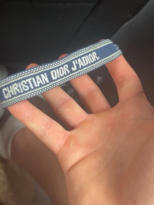 Blått Christian Dior J'Adior armband - Snyggt blått armband från Christian Dior med texten 'J'Adior' broderad i vitt. Armbandet är gjort i tyg och har en trendig, avslappnad look. Perfekt accessoar för att lyfta din outfit med en exklusiv touch.